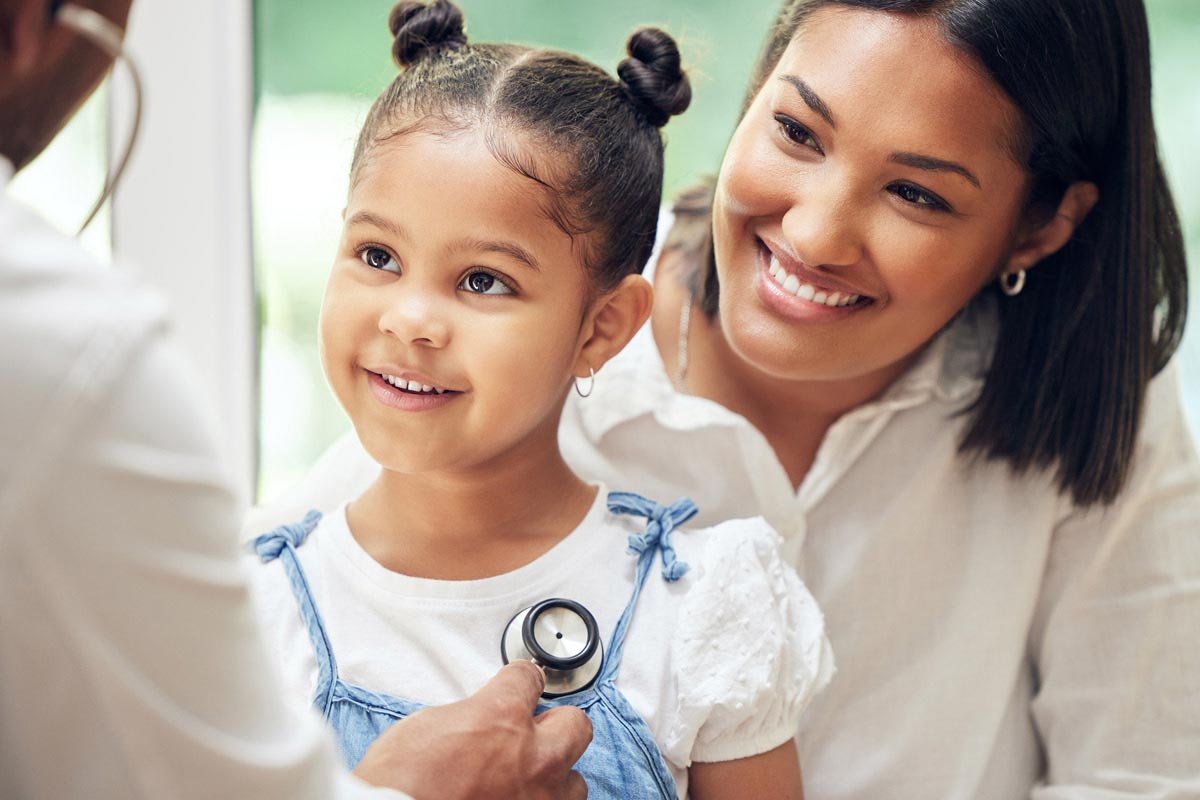 A doctor listens to a child's heartbeat using a stethoscope while her mother looks on with a smile.