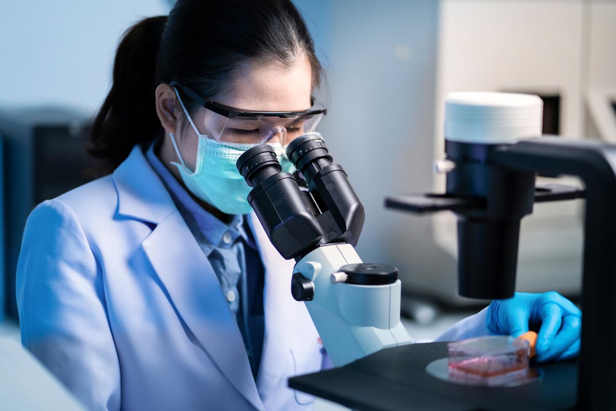 a female doctor wearing a white coat and face mask exams cells under a microscope in a lab