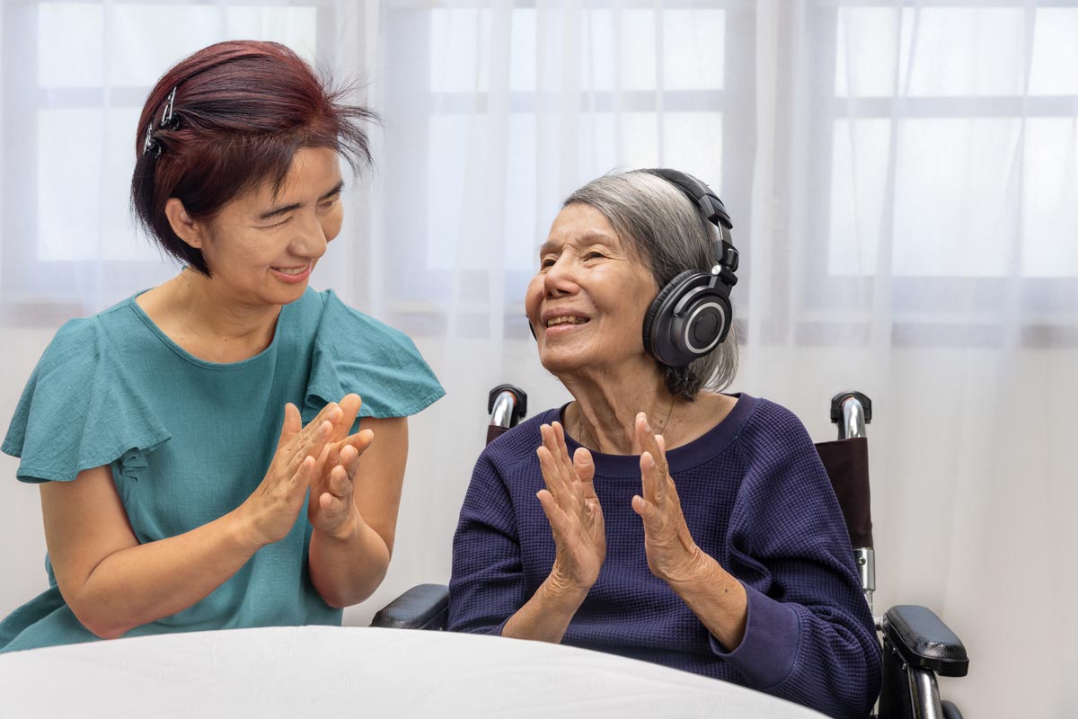 adult woman sits at a table and claps along with her elderly mother who listens to music on headphones