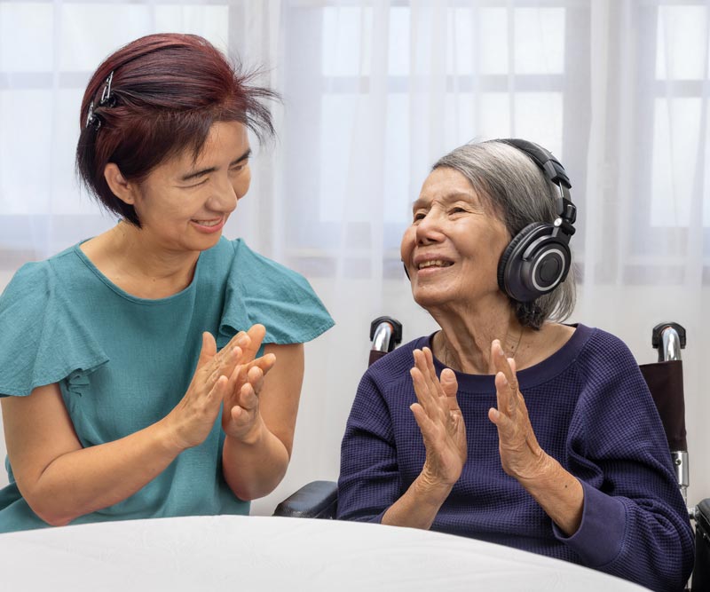 adult woman sits at a table and claps along with her elderly mother who listens to music on headphones