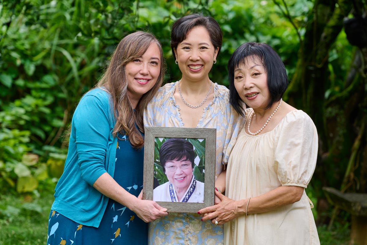 Three women stand close together holding a framed photo of another woman.