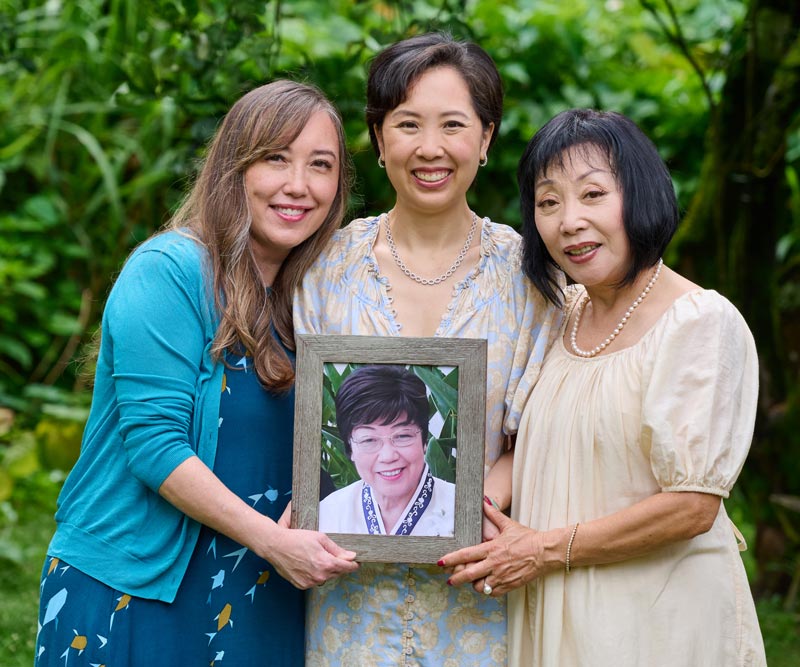 Three women stand close together holding a framed photo of another woman.