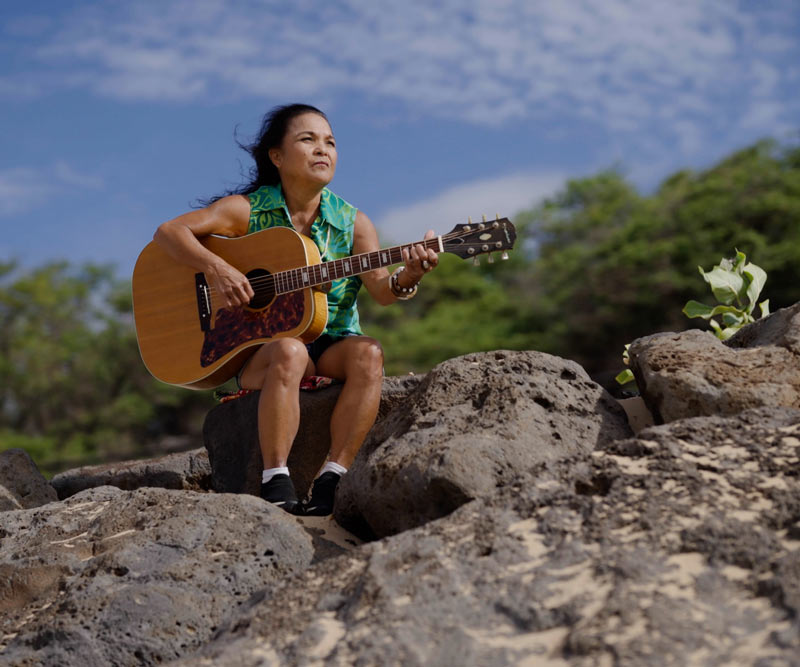 woman holds guitar while sitting on rocks on the side of a mountain