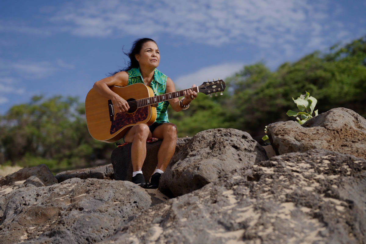 woman holds guitar while sitting on rocks on the side of a mountain