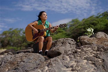 Woman holding a guitar sitting on a rocky outcropping and staring determinedly into the distance.