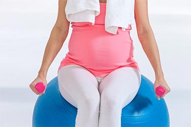 Pregnant woman sitting on exercise ball holding light weights in each hand.