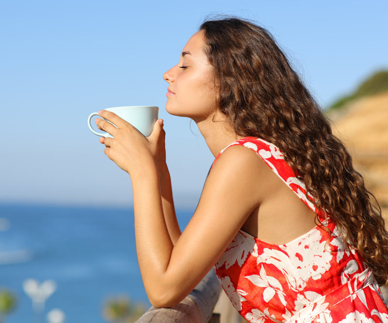 a woman holds a cup of tea and stands on a balcony overlooking the ocean