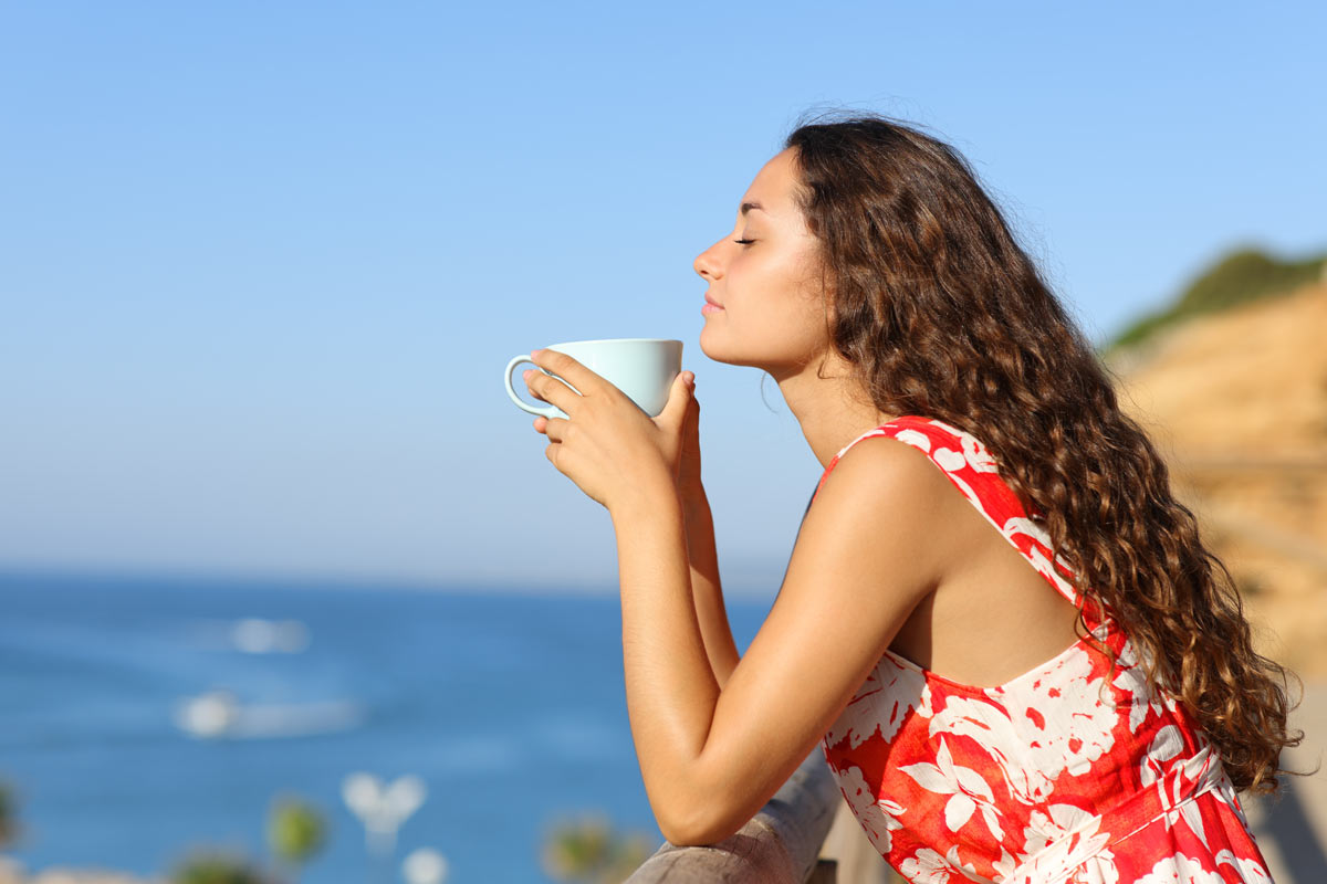 a woman holds a cup of tea and stands on a balcony overlooking the ocean