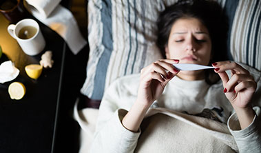 Woman sick in bed looking at a thermometer.