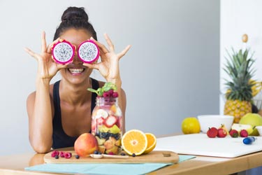 Smiling woman holds up dragon fruit halves over her eyes with a jar of fresh-cut fruit in front of her on a table