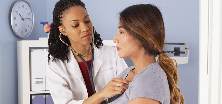 Woman doctor listens to heartbeat of woman patient in an exam room.