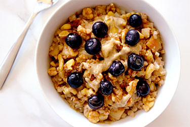 A bowl of Cauliflower Oats topped with blueberries sits on a marble counter top with a spoon resting in the background.