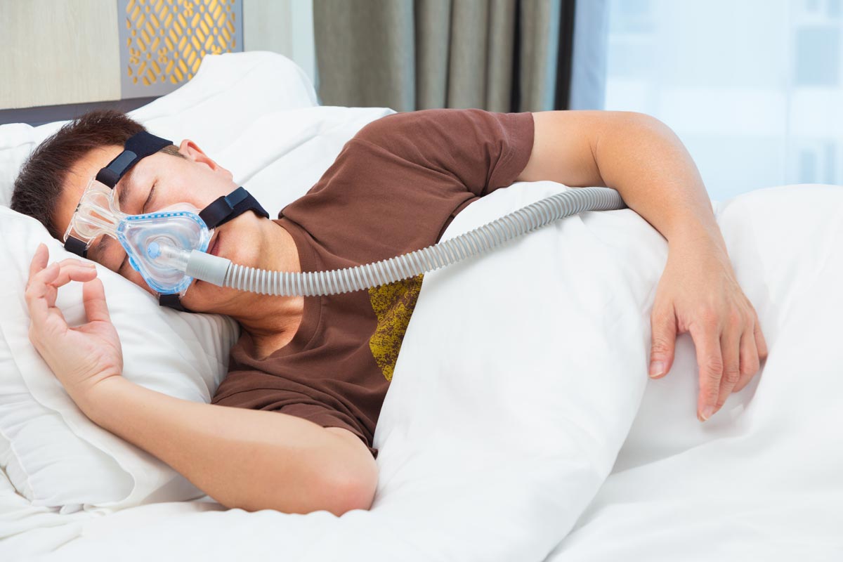 Asian man wearing CPAP mask, connecting to air hose, sleeping on his bed in his bedroom with large window