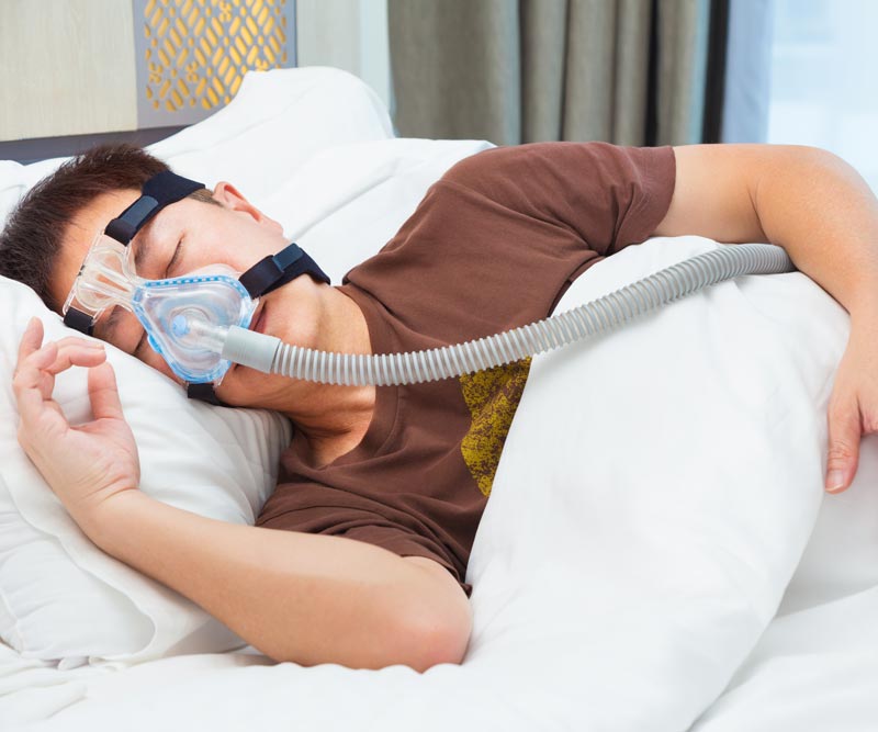 Asian man wearing CPAP mask, connecting to air hose, sleeping on his bed in his bedroom with large window