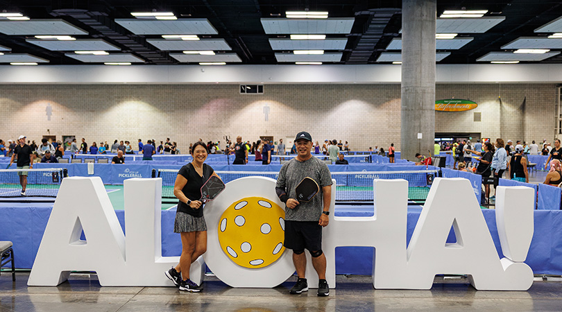 Two pickleball players next to a large ALOHA sign in front of some indoor pickleball courts.