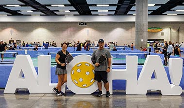 Pickleball players with paddles in front of courts and a giant Aloha sign.